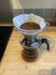 Image: V60 with ground coffee and filter paper sat on top of glass cafare on a kitchen counter.